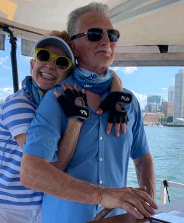 Happy mature couple on a harbor boat — woman in yellow sunglasses hugging man at the helm, both in blue shirts and sailing gloves with a waterfront city skyline and water in the background.