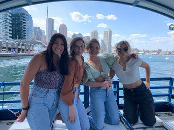 Four friends smiling and posing on a boat with a sunny downtown waterfront skyline and harbor in the background.