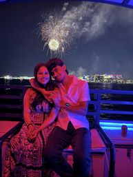 Couple on a boat watching fireworks over a city skyline at night, bathed in pink and blue lights reflecting on the water.