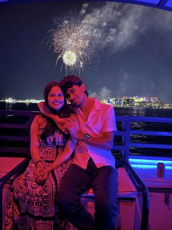 Couple on a boat watching fireworks over a city skyline at night, bathed in pink and blue lights reflecting on the water.