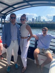 Three adults in summer boat attire and sunglasses on a covered harbor cruise with the Boston skyline and marina in the background.