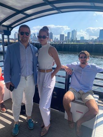 Three adults in summer boat attire and sunglasses on a covered harbor cruise with the Boston skyline and marina in the background.