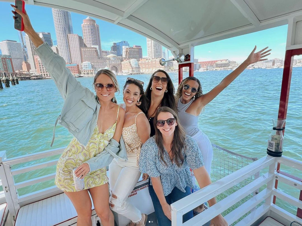 Five friends smiling and posing on a sunny boat in Boston Harbor with the downtown Boston skyline and waterfront in the background.