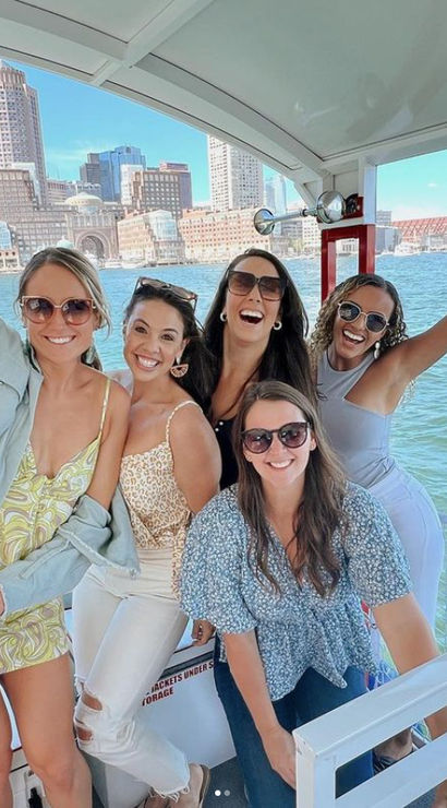 Five friends smiling and posing on a sunny boat in Boston Harbor with the downtown Boston skyline and waterfront in the background.