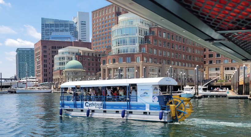 Pedal-powered tour boat with yellow paddlewheel cruising Boston Harbor in front of waterfront brick office buildings and moored yachts