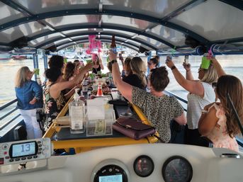 Group of women raising drinks and taking photos on a covered party boat at a marina, gathered around a central snack-and-drink bar with yachts and waterfront in the background.