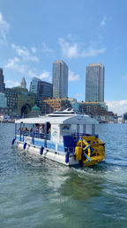 Pedal-powered tour boat with bright yellow paddlewheel cruising Boston Harbor in front of waterfront historic buildings and twin high-rise towers under a sunny blue sky.