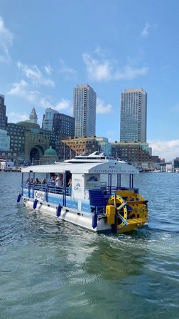 Pedal-powered tour boat with bright yellow paddlewheel cruising Boston Harbor in front of waterfront historic buildings and twin high-rise towers under a sunny blue sky.
