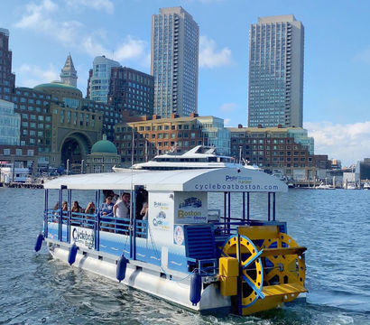 Pedal-powered tour boat with bright yellow paddlewheel cruising Boston Harbor in front of waterfront historic buildings and twin high-rise towers under a sunny blue sky.