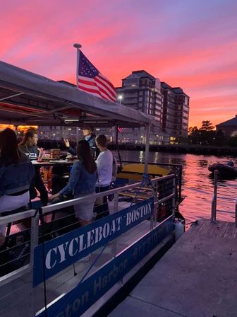 Group on a pedal-powered harbor tour boat at the Boston waterfront, American flag waving against a vivid pink sunset and city skyline.