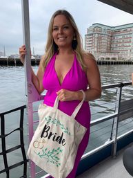 Person in a bright fuchsia halter dress holding a cream tote printed 'Bride' with rings, posing on a boat deck by a city harbor with waterfront buildings and cloudy sky — bride-to-be, bachelorette-on-the-water vibe.