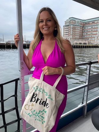 Person in a bright fuchsia halter dress holding a cream tote printed 'Bride' with rings, posing on a boat deck by a city harbor with waterfront buildings and cloudy sky — bride-to-be, bachelorette-on-the-water vibe.