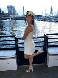 Smiling bride-to-be in a white mini dress and captain’s hat posing on a boat deck at an urban marina with sailboats and city skyline in the background