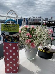 Pink carnations and baby's breath in a vase beside a polka-dot gift bag and captain's hat on a table by the Boston harbor skyline