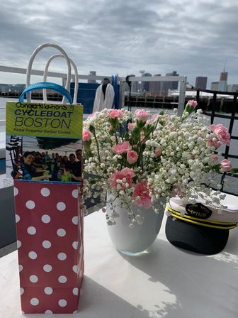 Pink carnations and baby's breath in a vase beside a polka-dot gift bag and captain's hat on a table by the Boston harbor skyline