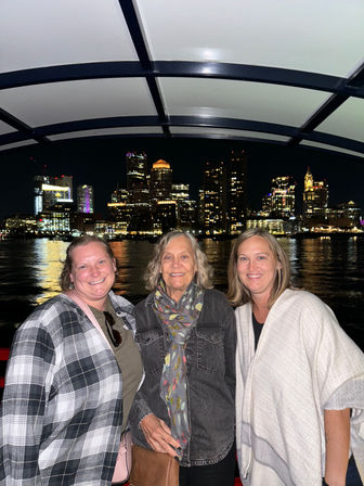 Three smiling women posing on a nighttime harbor boat with the Boston skyline and city lights reflecting on the water