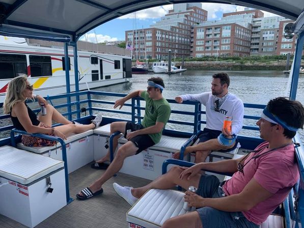 Four friends relaxing on a covered pontoon boat at an urban marina, sipping drinks with moored boats and brick waterfront apartments in the background.