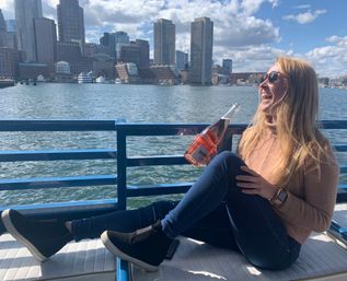 Laughing woman in sunglasses relaxing on a boat deck, holding a bottle of rosé with the Boston Harbor skyline and waterfront in the background on a sunny day.