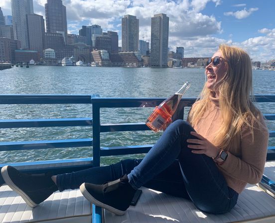 Laughing woman in sunglasses relaxing on a boat deck, holding a bottle of rosé with the Boston Harbor skyline and waterfront in the background on a sunny day.