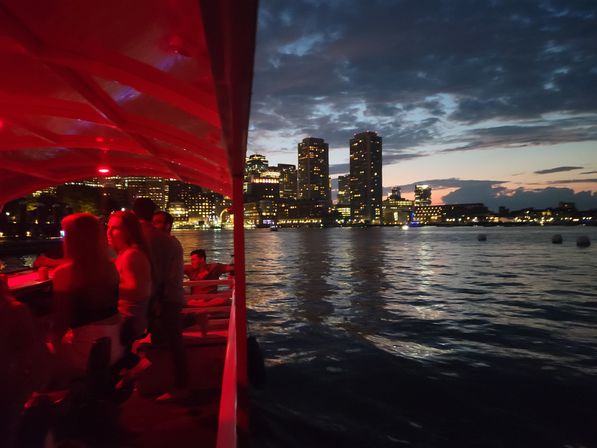 Passengers on a red-lit party boat at dusk with a glowing city skyline and high-rise lights reflecting on calm harbor water.