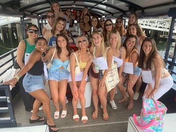 Smiling group of young women in summer outfits posing on a covered boat at a marina — casual waterfront boat cruise with drinks, tote bags, and a colorful beach bag.