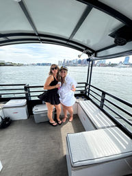 Two friends partying on a covered pontoon boat near a waterfront city skyline, one wearing a captain hat and sash, both holding drinks.
