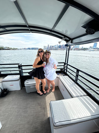 Two friends partying on a covered pontoon boat near a waterfront city skyline, one wearing a captain hat and sash, both holding drinks.