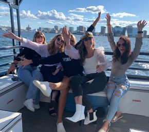 Five friends laughing and raising their arms on a sunny boat ride, wearing sunglasses and a captain's hat, with an urban waterfront skyline and sailboats across the water in the background.