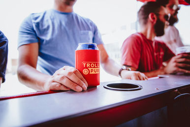 Sunlit close-up of a hand holding a canned beer in a red can cooler with a yellow logo on a bar counter, two casually dressed men chatting blurred in the background for a lively urban bar scene.