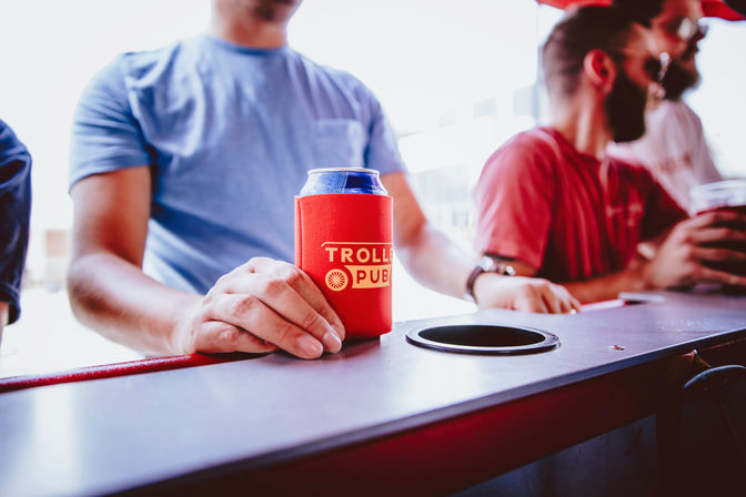 Sunlit close-up of a hand holding a canned beer in a red can cooler with a yellow logo on a bar counter, two casually dressed men chatting blurred in the background for a lively urban bar scene.
