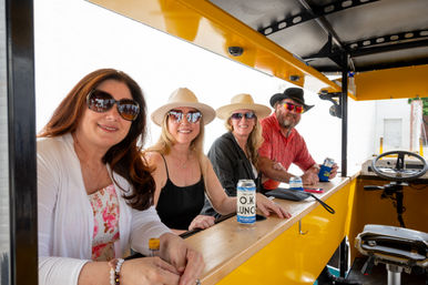 Four adults wearing sunglasses and hats smiling aboard a yellow pedal-pub bar, holding canned drinks on an outdoor group ride during a sunny pub-crawl style outing