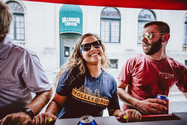 Smiling woman in sunglasses leaning at a red trolley-style bar with two friends holding canned drinks, enjoying a casual summer outing in a downtown streetscape with historic windows behind them.