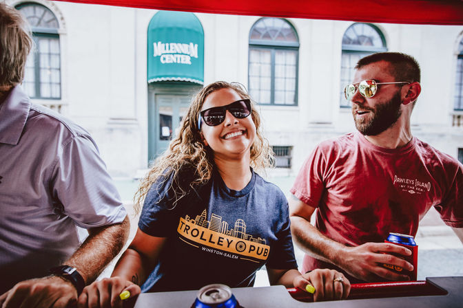 Smiling woman in sunglasses leaning at a red trolley-style bar with two friends holding canned drinks, enjoying a casual summer outing in a downtown streetscape with historic windows behind them.