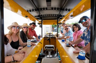 Eight adults wearing hats and sunglasses, smiling and holding canned drinks aboard a bright yellow pedal-powered party bike on a downtown street.
