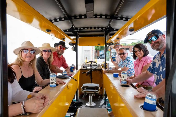 Eight adults wearing hats and sunglasses, smiling and holding canned drinks aboard a bright yellow pedal-powered party bike on a downtown street.