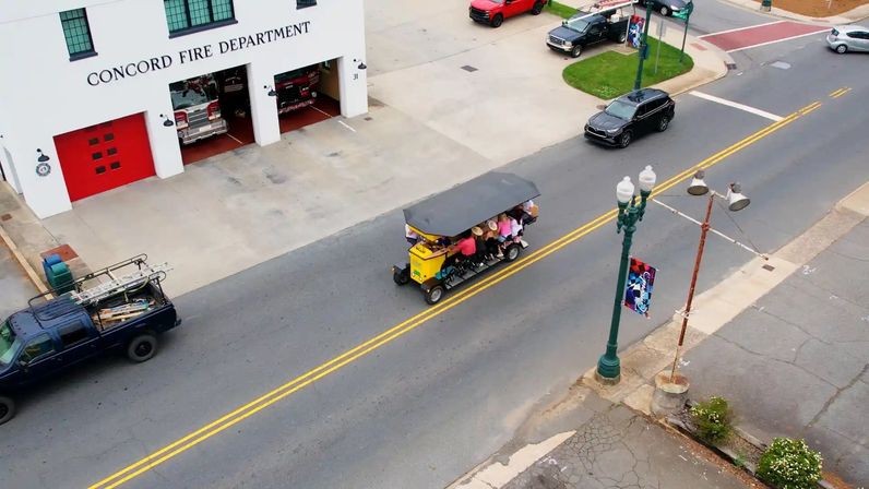 Aerial view of a yellow pedal-powered party bike full of riders rolling down a small-town main street past a white fire station with red doors and parked fire trucks