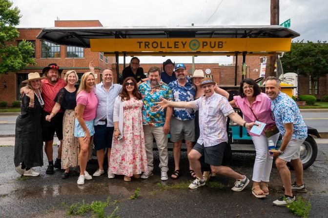 Group of smiling adults in colorful summer outfits and hats posing in front of a yellow open-air party trolley on a wet downtown street, lively group outing