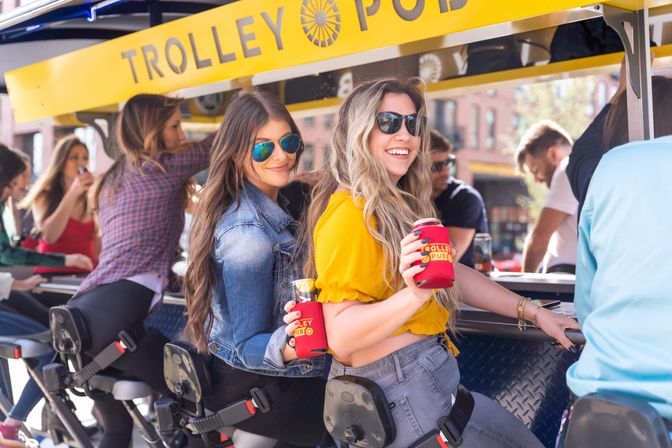 Smiling women in sunglasses sip canned drinks while seated at a sunny downtown pedal-trolley bar, friends enjoying a lively outdoor group outing.