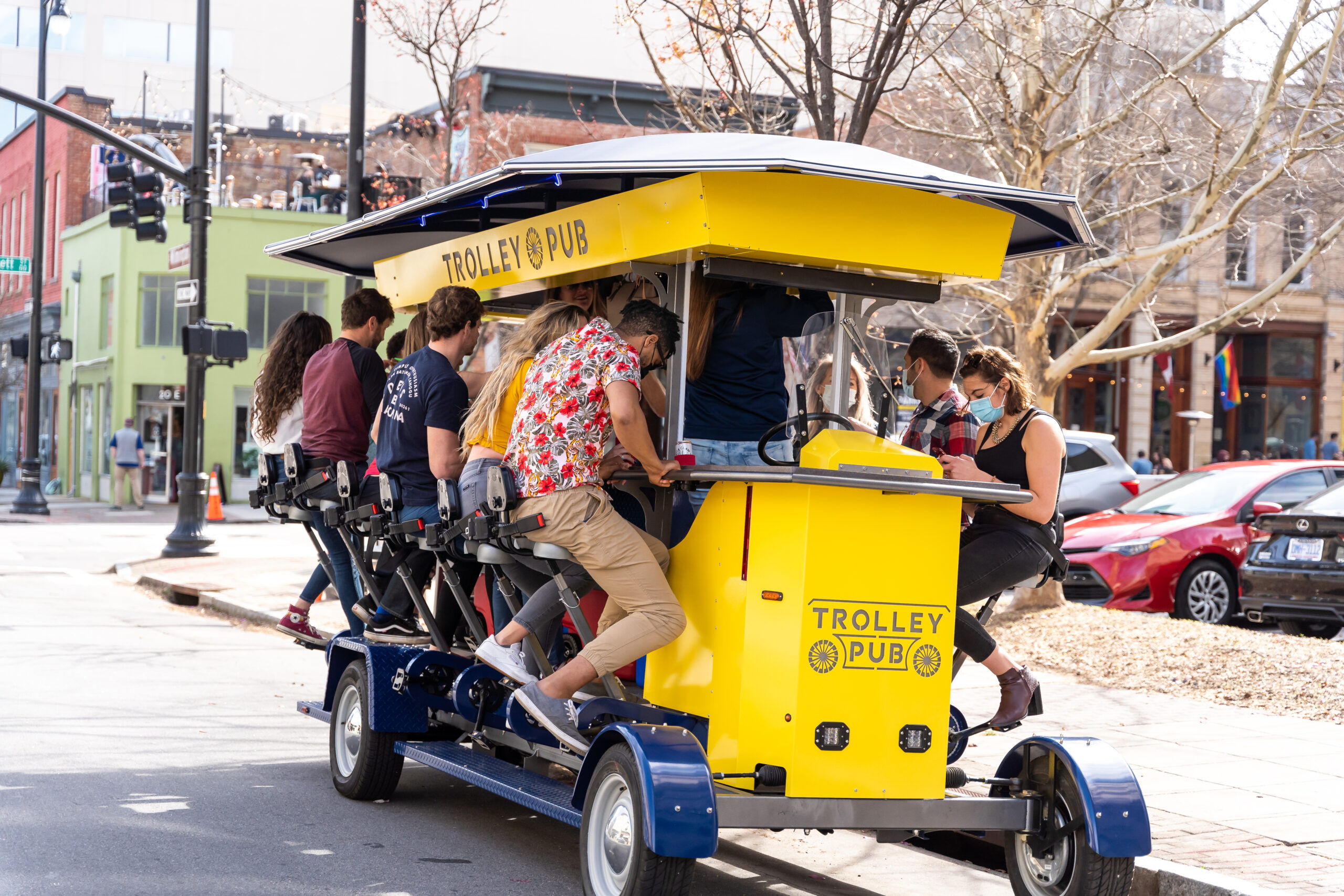 Yellow pedal-powered party trolley with a group seated at bar-style stations, pedaling and socializing on a downtown city street.
