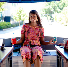 Smiling woman in a colorful jumpsuit seated on a sunny city party bike, holding a bottled drink in a koozie and a smartphone