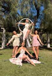 Bride in white dress holding a bouquet perched on an aerial hoop while three friends in pastel dresses pose on the grass beneath willow trees and a pergola in a sunny park