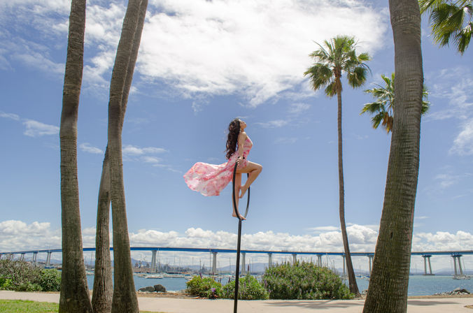Woman in a pink flowing dress perched on a hoop pole above a San Diego waterfront walkway, framed by tall palm trees and the Coronado Bridge over the bay
