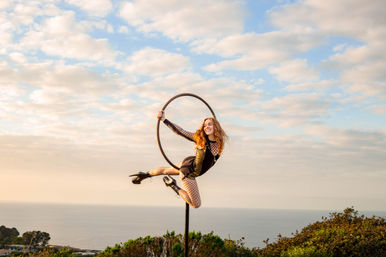 Aerial performer in a black costume posing on a suspended hoop above coastal cliffs, bathed in golden-hour light with a pastel cloudy sky and ocean horizon.
