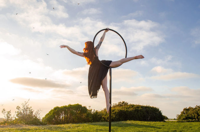 Aerial hoop performer in a black dress balances on a vertical hoop on a grassy hill at golden hour, red hair glowing against a pastel sky with birds.