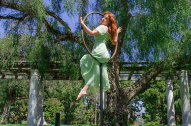 Aerial performer in a mint green dress posing on a hoop (lyra) suspended from a pergola in a sunlit park beneath a large willow tree