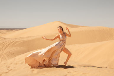 Barefoot dancer in a flowing champagne gown striking a graceful pose on sunlit golden sand dunes under a clear sky — dramatic desert photoshoot.