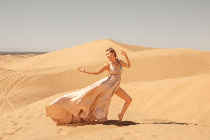 Barefoot dancer in a flowing champagne gown striking a graceful pose on sunlit golden sand dunes under a clear sky — dramatic desert photoshoot.