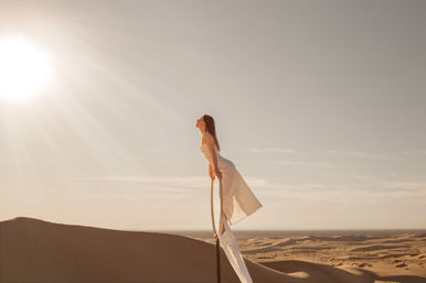 Woman in a flowing white dress balancing on an aerial hoop above golden sand dunes under a bright sun at golden hour — serene desert aerial scene