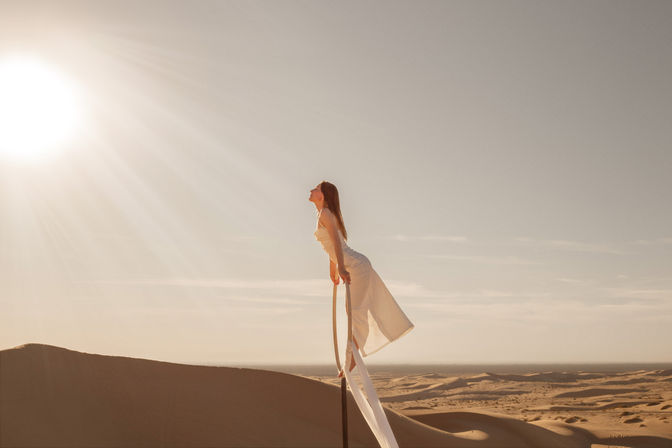 Woman in a flowing white dress balancing on an aerial hoop above golden sand dunes under a bright sun at golden hour — serene desert aerial scene
