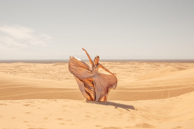 Sunlit desert dunes with a person in a billowing silk gown striking a dancer-like pose, flowing fabric against a wide sandy landscape and pale blue sky.